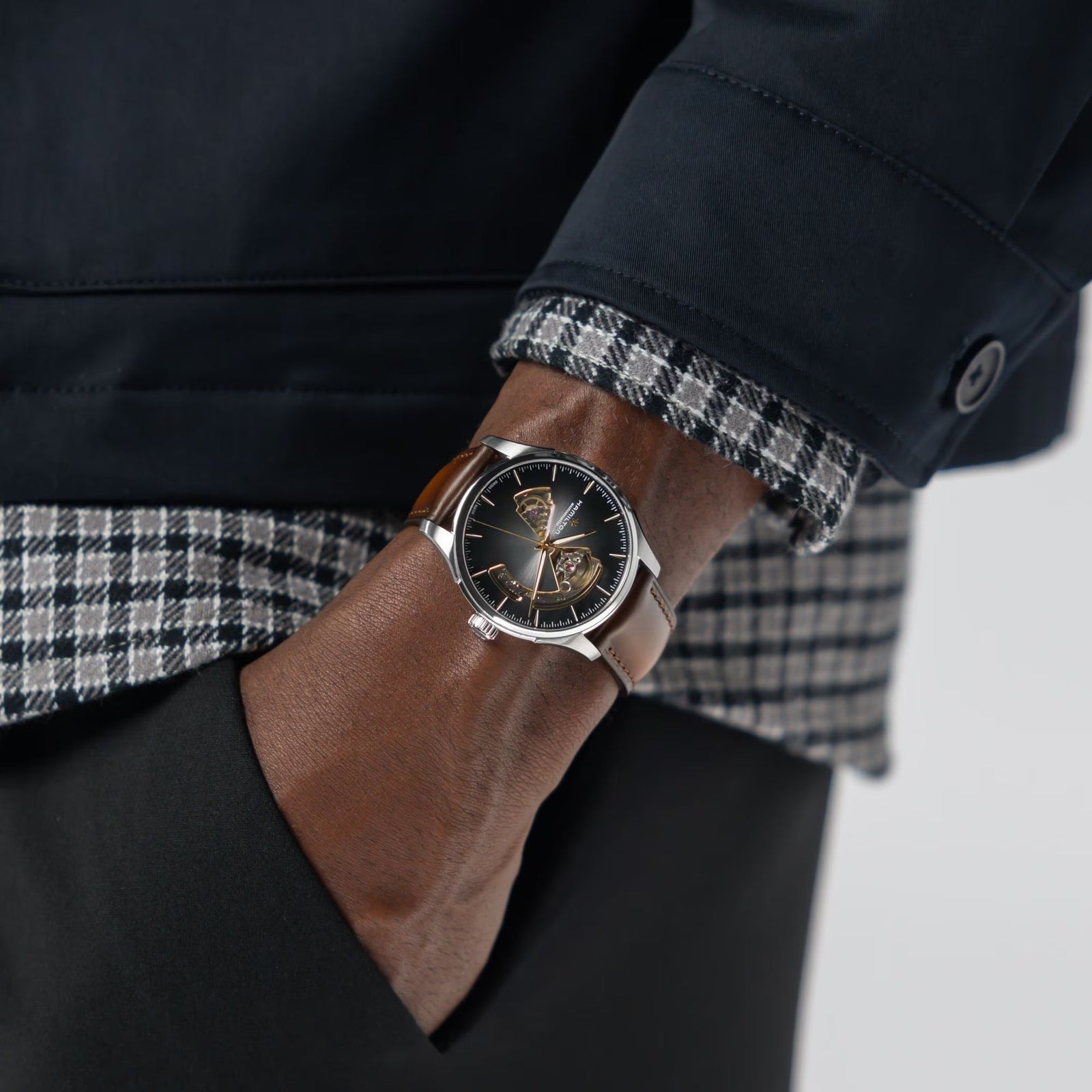 Person wearing a watch with a dark face and brown strap, dressed in a dark coat and plaid shirt.
Hamilton Jazzmaster Open Heart Watch H32675501 Bandiera Jewellers